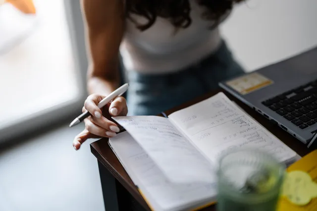 A person is seated at a desk with a laptop open in front. They hold a pen in one hand and flip through a notebook with notes. A glass of water is nearby. Sunlight is coming through a window. Interview prompts for finance audiences that never invite an advice request