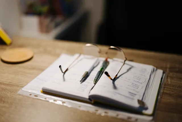 A person is studying at home during the evening. A notebook lies open on the desk with several pens. Reading glasses rest on the notebook, indicating active use. A ninety-day finance creator pilot that ends with a decision, not a launch