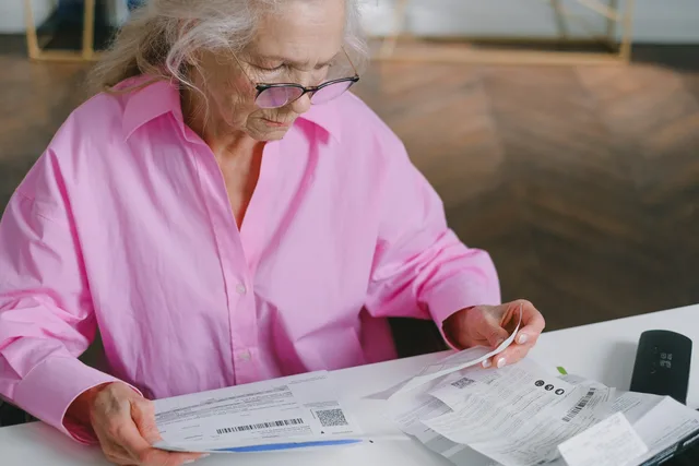 Elderly woman in pink blouse reading documents at a table indoors. FIRE YouTube UK sponsor-brief checklist: ISA, pension and FCA checks